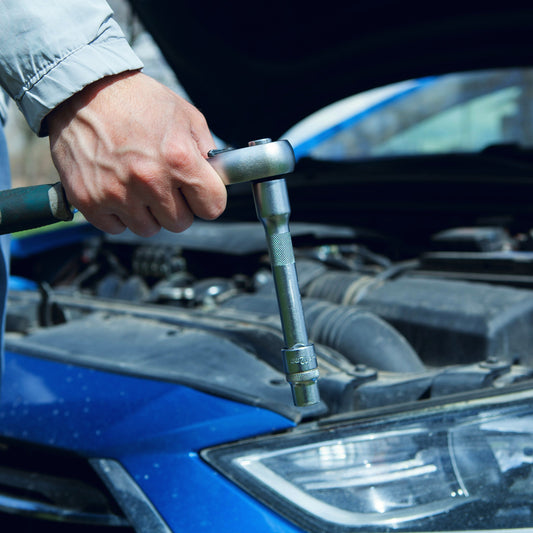 A hand turning a T-bar socket wrench on a car engine, bonnet open over a blue car